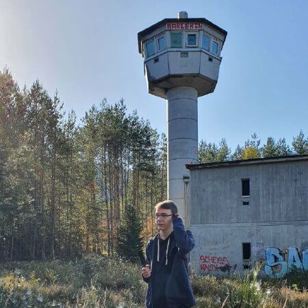 Ein Mann steht vor einem alten Wachturm und einem verlassenen Gebäude in Ludwigswinkel, umgeben von Bäumen und Graffiti.