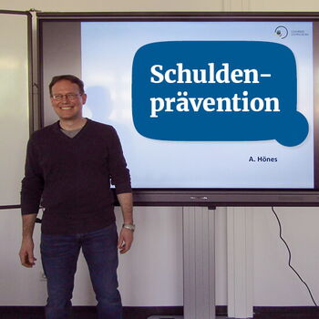 Ein Mann steht lächelnd neben einem Bildschirm, der das Thema "Schuldenprävention" präsentiert.
