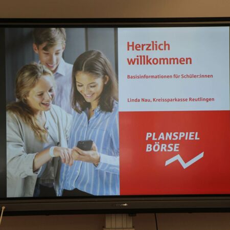 Drei Schüler schauen lächelnd auf ein Smartphone, während auf einem Bildschirm Informationen zum Planspiel Börse angezeigt werden.