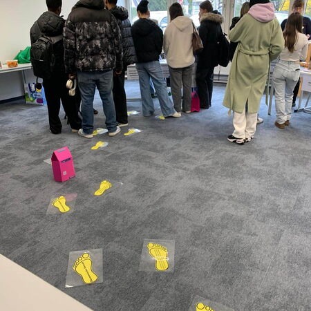 Eine Gruppe von Menschen wartet in einem Raum, während gelbe Fußabdrücke den Weg zu einem Stand weisen.