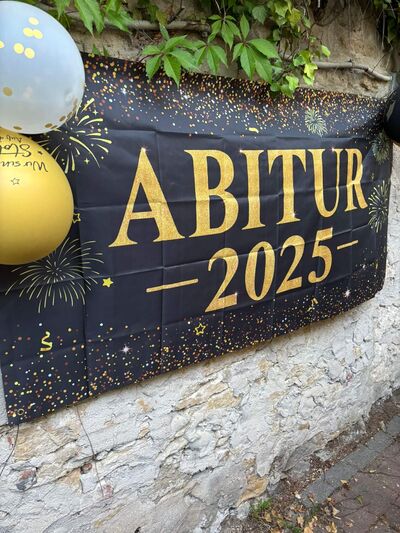Ein Banner mit der Aufschrift "Abitur 2025" hängt an einer Wand, dekoriert mit bunten Luftballons und Feuerwerksmotiven.