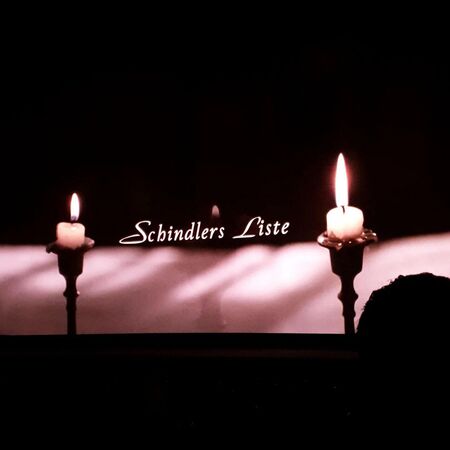 Zwei brennende Kerzen flankieren den Titel "Schindlers Liste" auf einem dunklen Hintergrund, der eine ernste Stimmung erzeugt.