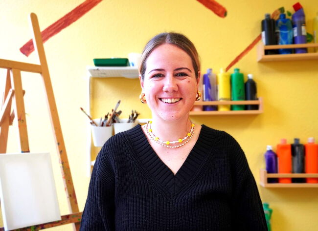 Eine lächelnde Frau steht in einem farbenfrohen Kunstzimmer der Grundschule am Flugfeld vor einem Regal mit Malutensilien.