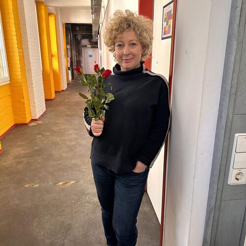 Eine Frau mit lockigem Haar hält einen Strauß roter Rosen in einem hellem Flur mit orangefarbenen Wänden.