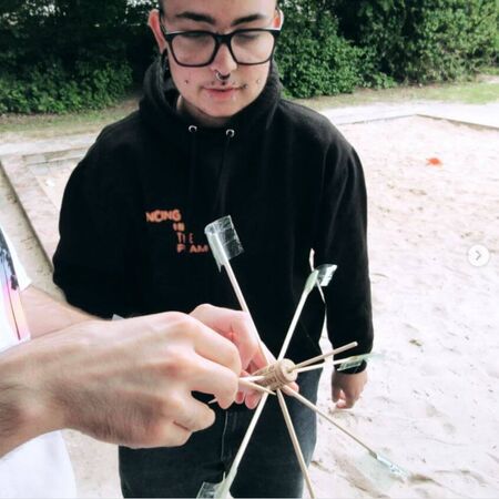 Eine Person im schwarzen Hoodie hält ein selbstgebautes Spielgerät aus Korken und Stäben auf einem Sandspielplatz.