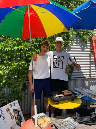 Zwei Männer grillen fröhlich Würstchen unter einem bunten Sonnenschirm im Garten.