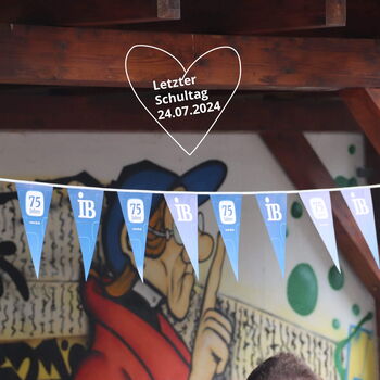 Blaue Wimpel mit "75 Jahre IB" hängen vor einer Wandmalerei, während ein Herz den letzten Schultag am 24.07.2024 ankündigt.