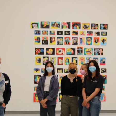 Vier maskierte Personen stehen vor einer bunten Kunstinstallation mit abstrakten Herzstücken an einer weißen Wand.