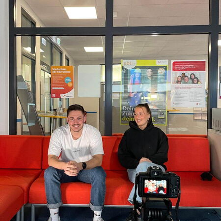 Zwei Personen sitzen auf einem roten Sofa und werden von einer Kamera gefilmt, als ob sie ein Interview führen.