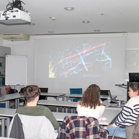 Personen sitzen in einem Seminarraum und schauen sich eine Präsentation an, die auf eine Leinwand projiziert wird.