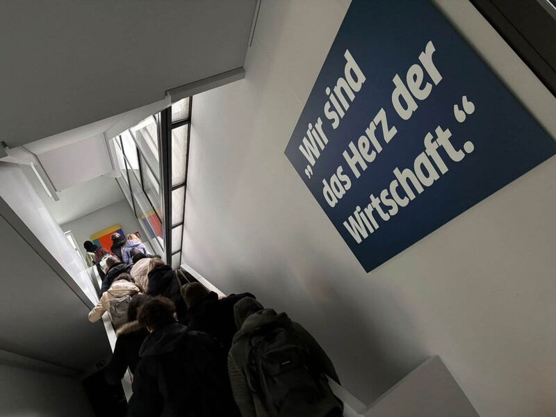 Menschen steigen in einem Gebäude eine Treppe hinauf, neben einem Schild mit der Aufschrift „Wir sind das Herz der Wirtschaft“.