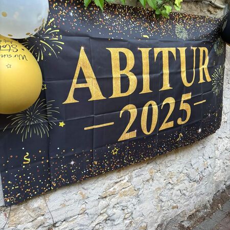 Ein Banner mit der Aufschrift "Abitur 2025" hängt an einer Wand, dekoriert mit bunten Luftballons und Feuerwerksmotiven.