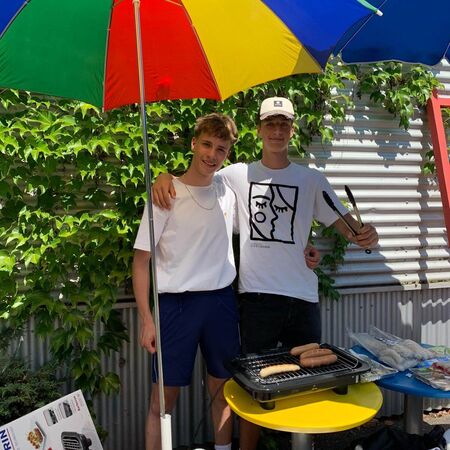 Zwei Männer grillen fröhlich Würstchen unter einem bunten Sonnenschirm im Garten.