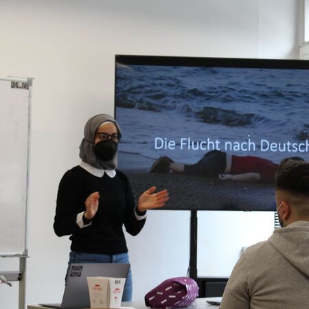 Eine Frau hält eine Präsentation über "Die Flucht nach Deutschland" vor einem großen Bildschirm in einem Klassenzimmer.