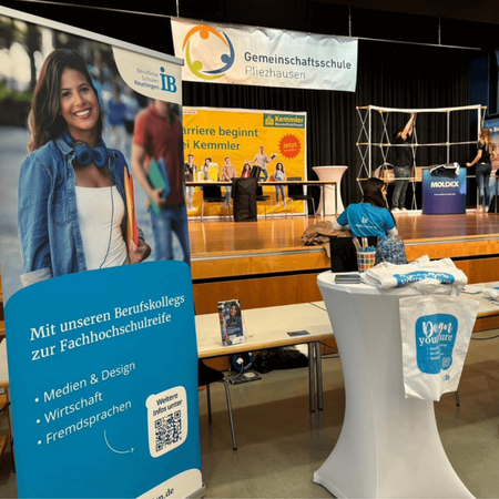 Ein Messestand in Pliezhausen zeigt Bildungsangebote und T-Shirts, während im Hintergrund ein Banner der Gemeinschaftsschule hängt.