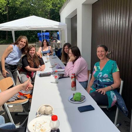Eine Gruppe von Menschen sitzt fröhlich an einem langen Tisch im Freien unter einem Pavillon und genießt das sonnige Wetter.