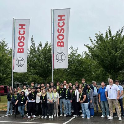 Eine Gruppe von Menschen steht lächelnd vor Bosch-Flaggen auf einem Parkplatz, umgeben von grünen Bäumen.
