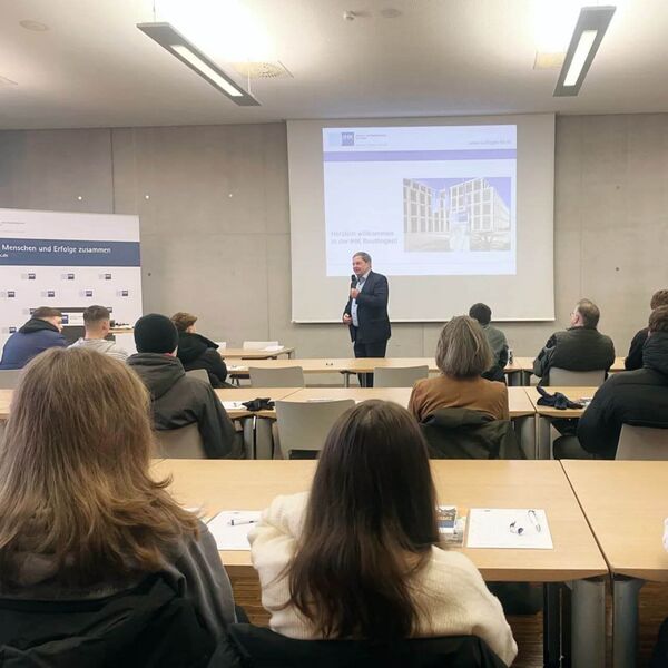 Ein Vortragender spricht vor einer Gruppe von Zuhörern in einem Konferenzraum, während eine Präsentation auf der Leinwand angezeigt wird.