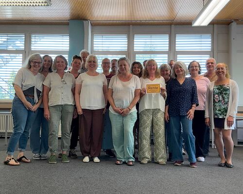 Eine Gruppe von Frauen hält ein Schild mit der Aufschrift „Rente“ in einem hellen Raum mit Fenstern.