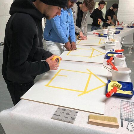 Fünf Personen arbeiten konzentriert an Tafel mit gelbem Klebeband, um geometrische Muster für ein kreatives Projekt zu erstellen.
