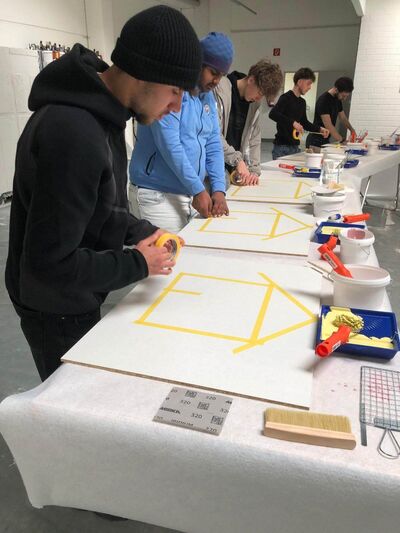 Fünf Personen arbeiten konzentriert an Tafel mit gelbem Klebeband, um geometrische Muster für ein kreatives Projekt zu erstellen.