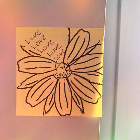 Ein gelber Zettel mit einer gezeichneten Blume und dem Wort "Love" mehrfach darauf steht an einer Wand.