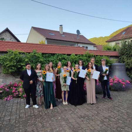 Eine Gruppe junger Menschen posiert draußen mit Urkunden und Sonnenblumen für ein Abschlussfoto.