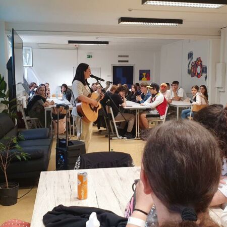 Eine Frau spielt Gitarre und singt vor einer aufmerksam zuhörenden Gruppe von jungen Erwachsenen in einem hellen Raum.