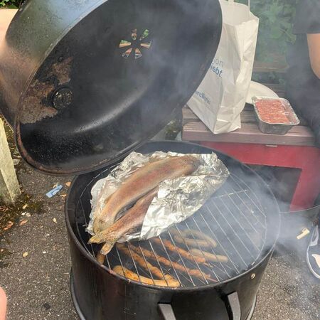 Frische Forellen werden in Alufolie auf einem runden Grill im Freien geräuchert, während Rauch aufsteigt.