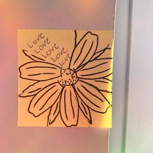 Ein gelber Zettel mit einer gezeichneten Blume und dem Wort "Love" mehrfach darauf steht an einer Wand.