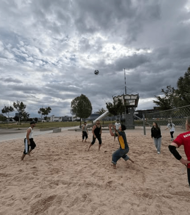 Eine Gruppe von Menschen spielt Volleyball im Sand unter einem bewölkten Himmel während des Projekttags.