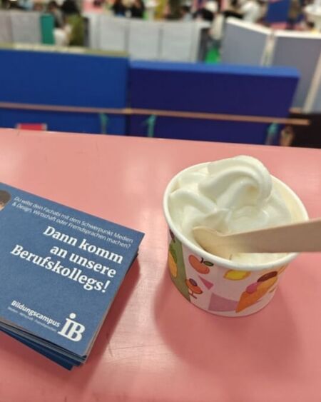 Ein Becher Eiscreme und Flyer für Berufskollegs liegen auf einem Tisch im lebendigen Messeumfeld.