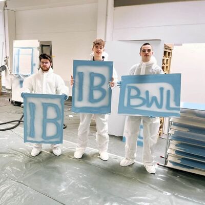 Drei Personen in Schutzanzügen halten Schilder mit den Buchstaben "IB" und "BWL" in einer Werkstatt, die Jobrotation darstellt.