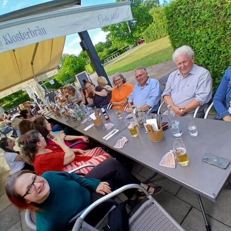 Eine Gruppe fröhlicher Menschen genießt Getränke und Gespräche unter einem großen Sonnenschirm im Freien.