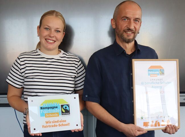 Zwei Personen halten Urkunden für die Fairtrade-Schule Asperg, strahlend in einem Schulraum.