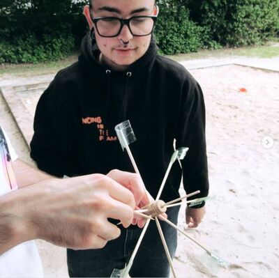 Eine Person im schwarzen Hoodie hält ein selbstgebautes Spielgerät aus Korken und Stäben auf einem Sandspielplatz.