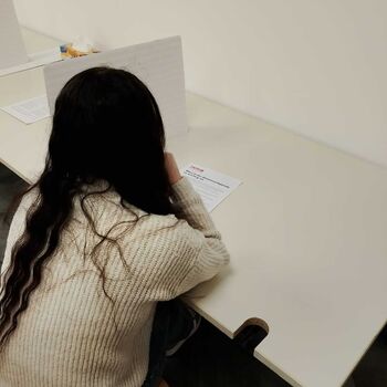Ein Mädchen mit langen Haaren sitzt an einem Tisch, konzentriert auf ein Blatt Papier vor sich.