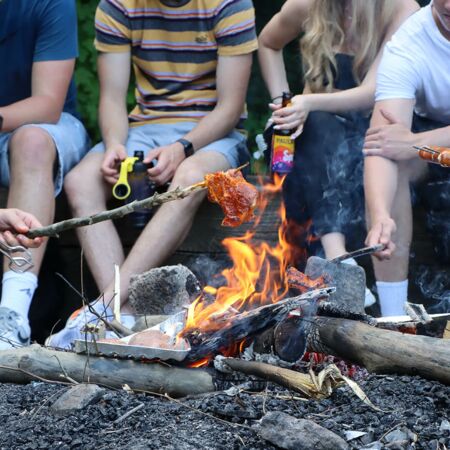Junge Menschen sitzen um ein Lagerfeuer und grillen Würstchen und Fleisch über den Flammen.