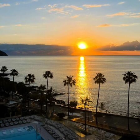 Die Sonne geht über dem Meer auf Mallorca unter, während Palmen und ein leerer Pool die friedliche Küstenszene rahmen.