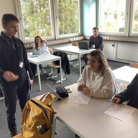 Ein Mann spricht in einem Klassenzimmer mit zwei lachenden Studierenden, während andere im Hintergrund arbeiten.