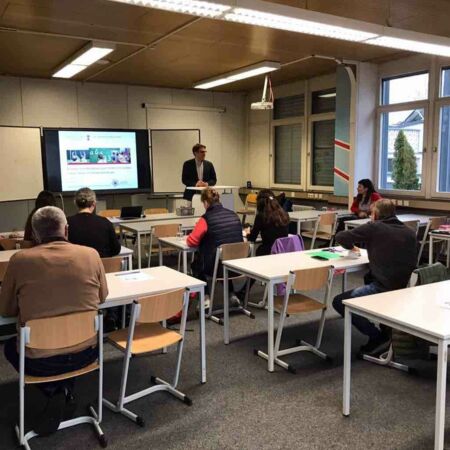 In einem Klassenzimmer hält ein Lehrer eine Präsentation vor einer Gruppe aufmerksam lauschender Erwachsener.