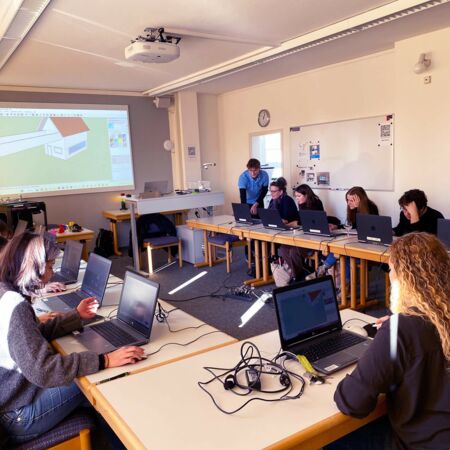 Eine Gruppe von Studierenden arbeitet an Laptops in einem Klassenraum, während ein Architekturmodell projiziert wird.