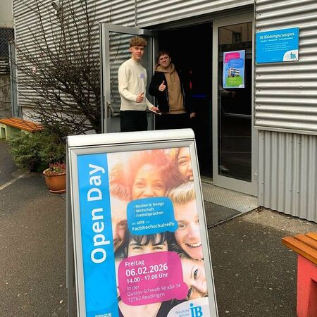 Zwei junge Männer stehen lächelnd am Eingang eines Gebäudes mit einem Plakat für den Tag der offenen Tür am 06.02.2026.
