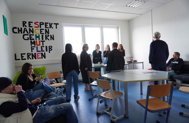 In einem Schulraum stehen Schüler und Lehrer zusammen, während an der Wand inspirierende Begriffe wie „RESPEKT“ und „CHANCEN“ hängen.