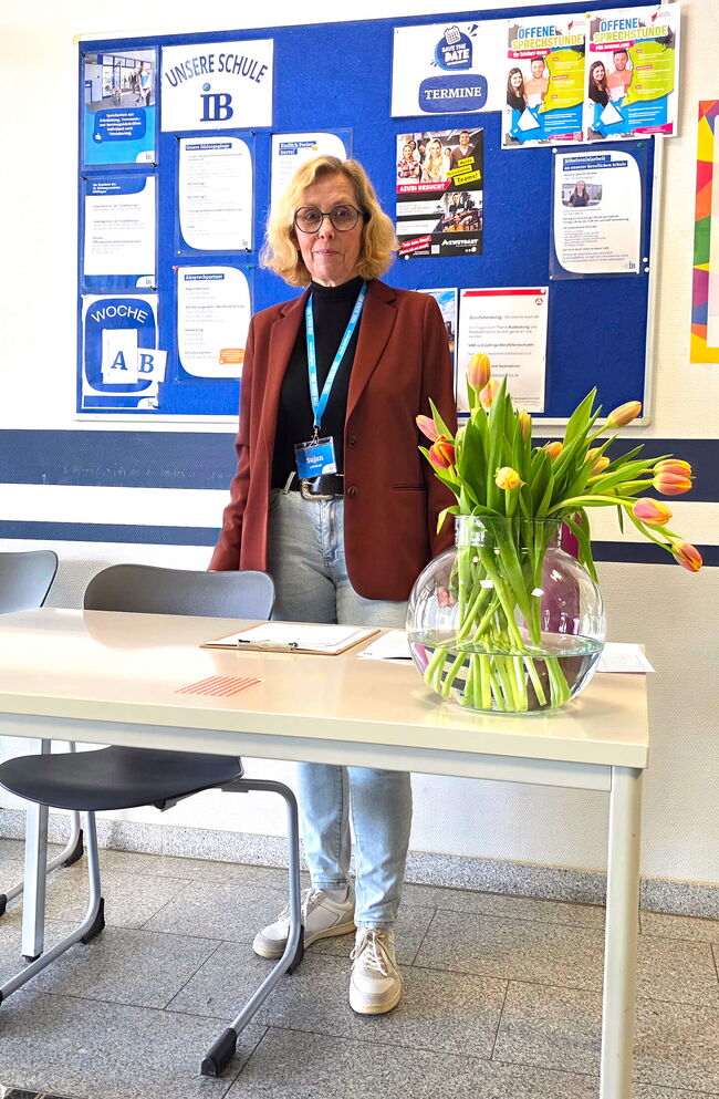 Eine Lehrkraft steht an einem Tisch mit einer Vase voll Tulpen, während im Hintergrund Informationen zur Schule präsentiert werden.