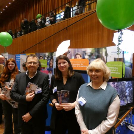 Drei Personen stehen lächelnd an einem Informationsstand mit Broschüren und grünen Luftballons in einer Messehalle.
