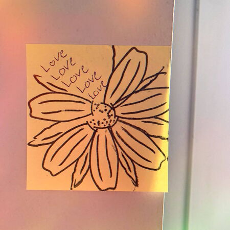 Ein gelber Zettel mit einer gezeichneten Blume und dem Wort "Love" mehrfach darauf steht an einer Wand.
