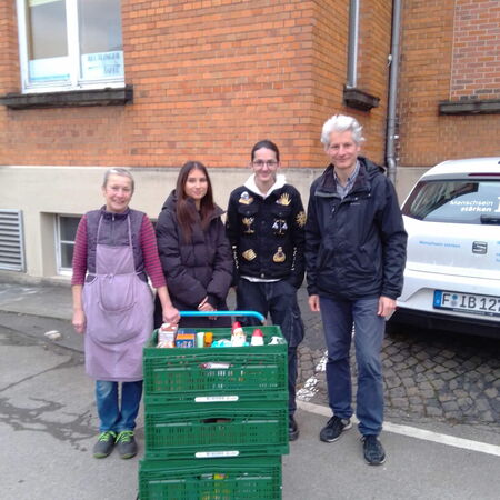 Vier Personen stehen neben einem mit Lebensmitteln beladenen Wagen vor einem Backsteingebäude bei einer Adventsspendaktion.