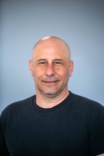 Ein Mann in einem schwarzen T-Shirt lächelt vor einem neutralen, grauen Hintergrund.
