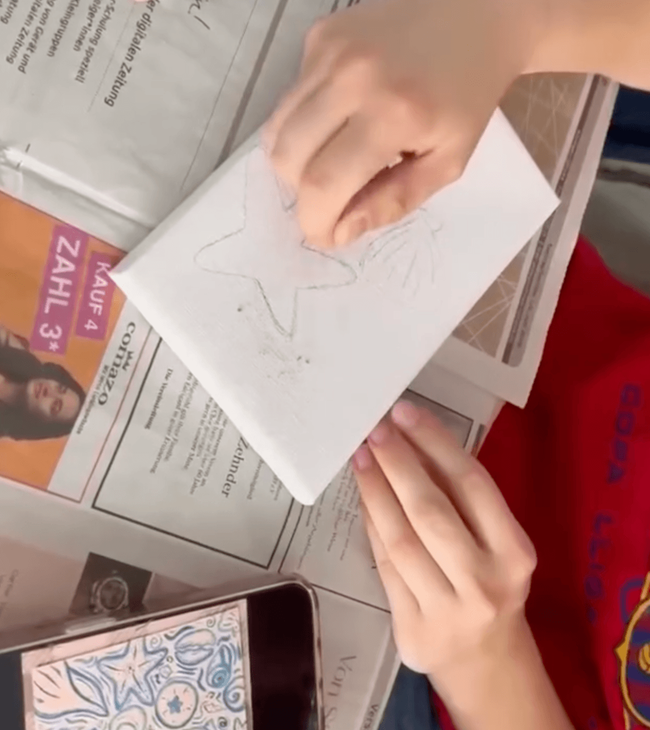Ein Kind zeichnet mit einem Bleistift auf einem Blatt Papier einen Stern, während es auf einer Zeitung sitzt.